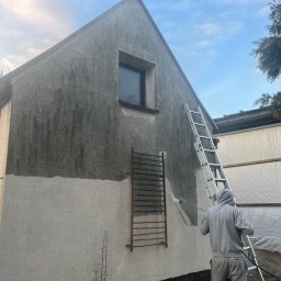 Dominik Parniak - Czyszczenie elewacji domu z szarego tynku za pomocą myjki ciśnieniowej, widoczna drabina i osoba w szarej bluzie z kapturem, część elewacji już umyta.