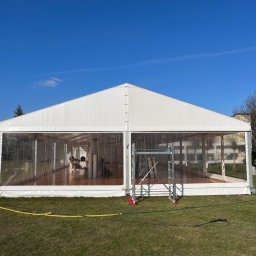 Dominik Parniak - Biały namiot imprezowy z przezroczystymi ścianami bocznymi, ustawiony na trawniku w słoneczny dzień, widoczne rusztowanie i żółty wąż doprowadzający wodę.