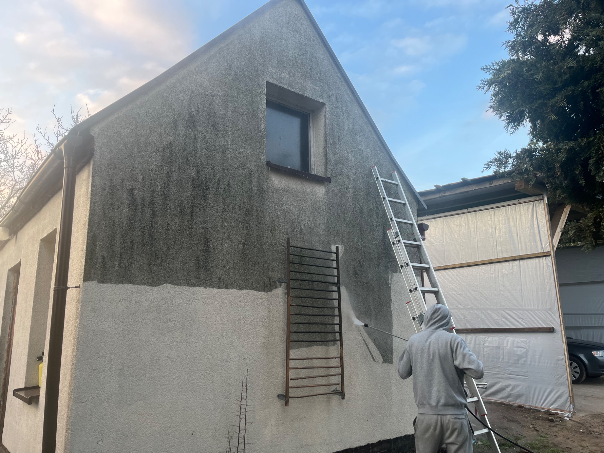 Czyszczenie elewacji domu z szarego tynku za pomocą myjki ciśnieniowej, widoczna drabina i osoba w szarej bluzie z kapturem, część elewacji już umyta.