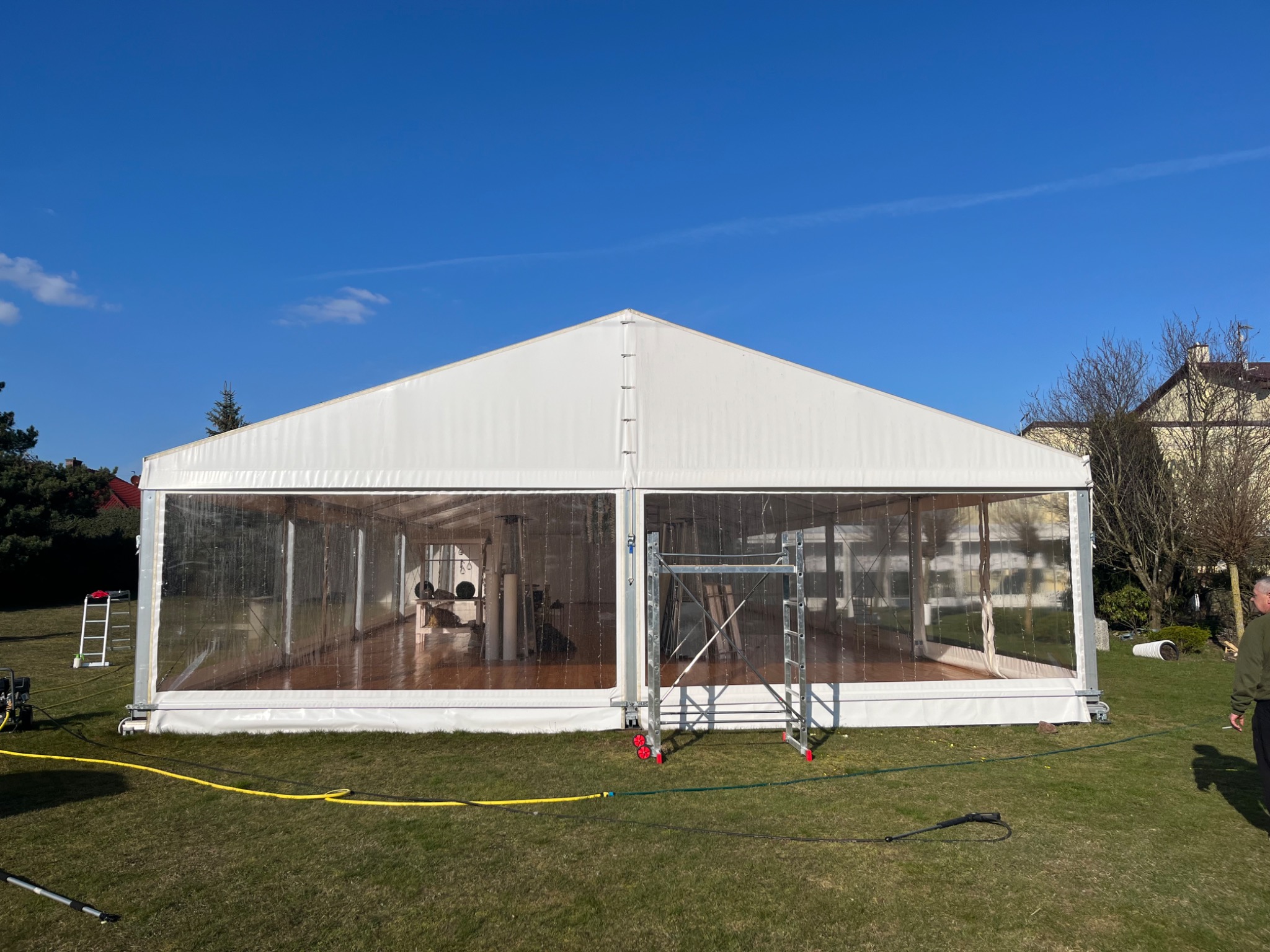 Biały namiot imprezowy z przezroczystymi ścianami bocznymi, ustawiony na trawniku w słoneczny dzień, widoczne rusztowanie i żółty wąż doprowadzający wodę.