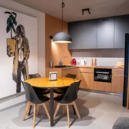 Nowoczesna kuchnia z drewnianymi i szarymi szafkami, okrągłym stołem z czarnymi krzesłami i dużym obrazem na ścianie.