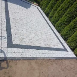 Ułożona jasnoszara kostka brukowa z ciemnoszarą obwódką, widok z góry na tle rzędu tuj i ziemi.