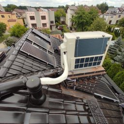 KLIWENT - Widok z góry na instalację pompy ciepła na dachu domu z czarną dachówką i panelami słonecznymi, w tle zabudowa jednorodzinna i zieleń.
