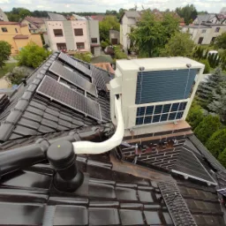 Widok z góry na instalację pompy ciepła na dachu domu z czarną dachówką i panelami słonecznymi, w tle zabudowa jednorodzinna i zieleń.