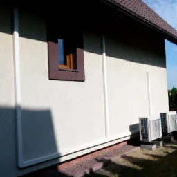 Dwa zewnętrzne moduły klimatyzacji zamontowane na betonowych podstawach przy ścianie domu z brązowym dachem, widoczne elementy instalacji odprowadzającej skropliny.