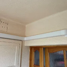 Biały kanał wentylacyjny poprowadzony pod sufitem w rogu pomieszczenia, widoczny na tle ściany o fakturze baranka i drewnianych drzwi z szybą.