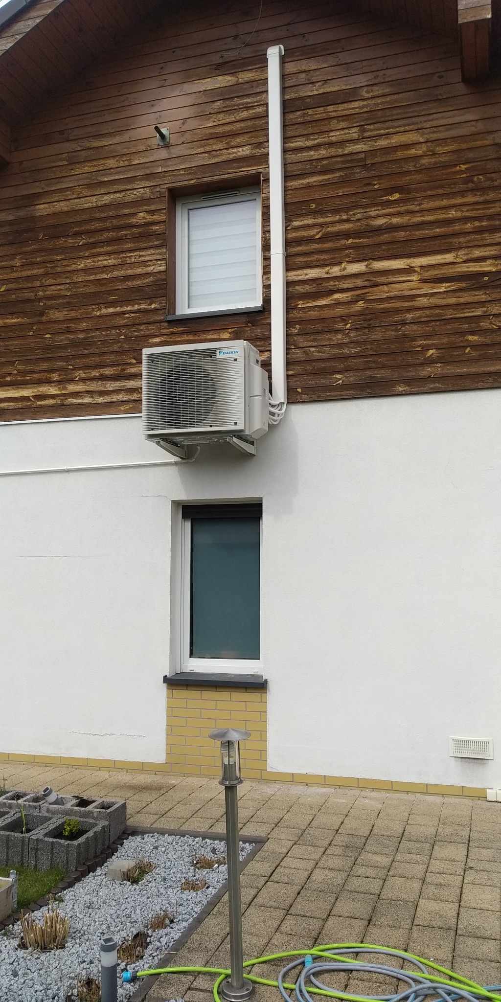 Zewnętrzna jednostka klimatyzacji Daikin zamontowana na ścianie domu z drewnianą elewacją powyżej białej, otynkowanej części, widoczna rura odprowadzająca skropliny.