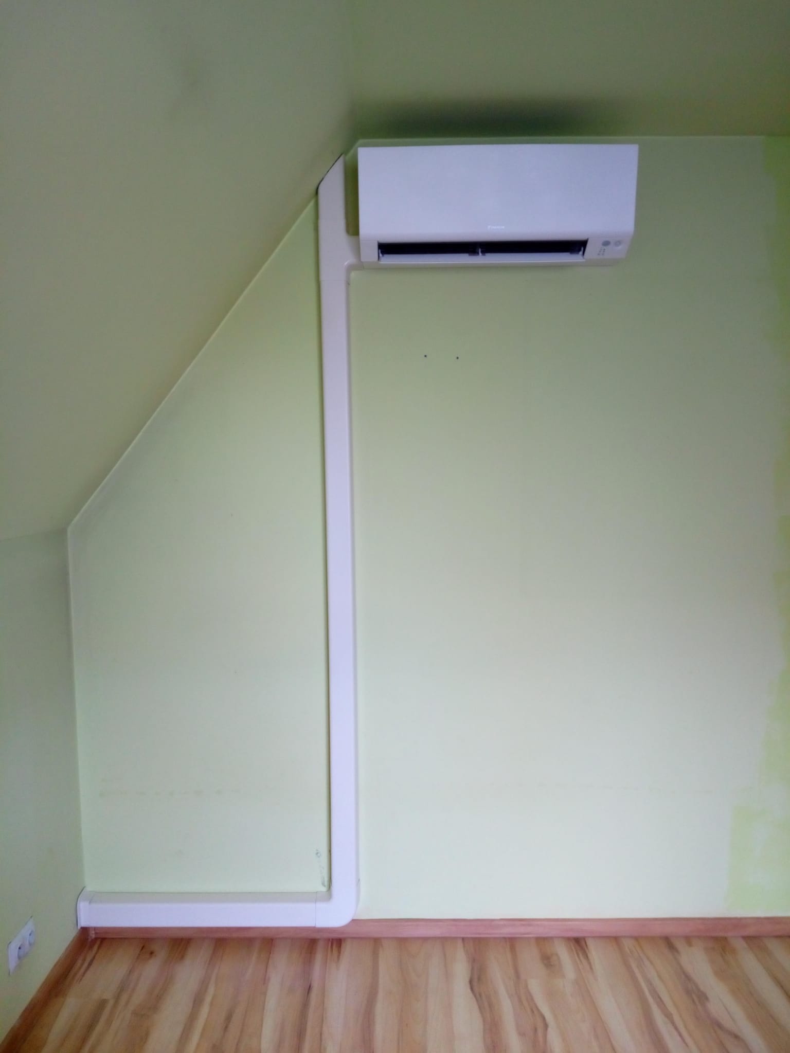 Biały klimatyzator na zielonej ścianie poddasza, z widocznymi rurami instalacyjnymi w białej listwie przypodłogowej i panelami podłogowymi.