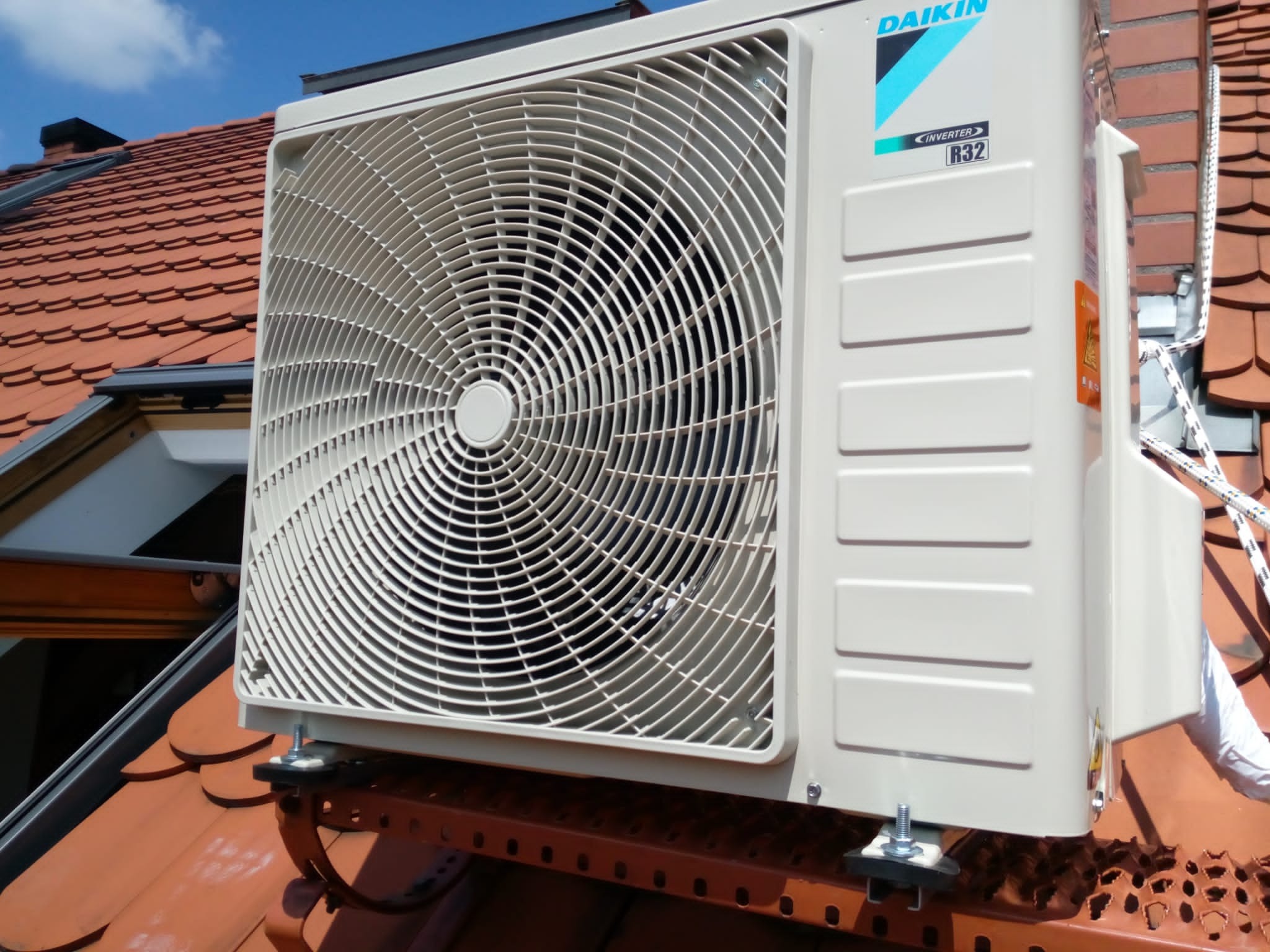Zewnętrzna jednostka klimatyzacji Daikin zamontowana na dachu z czerwonej dachówki, widoczne mocowania i logo producenta.