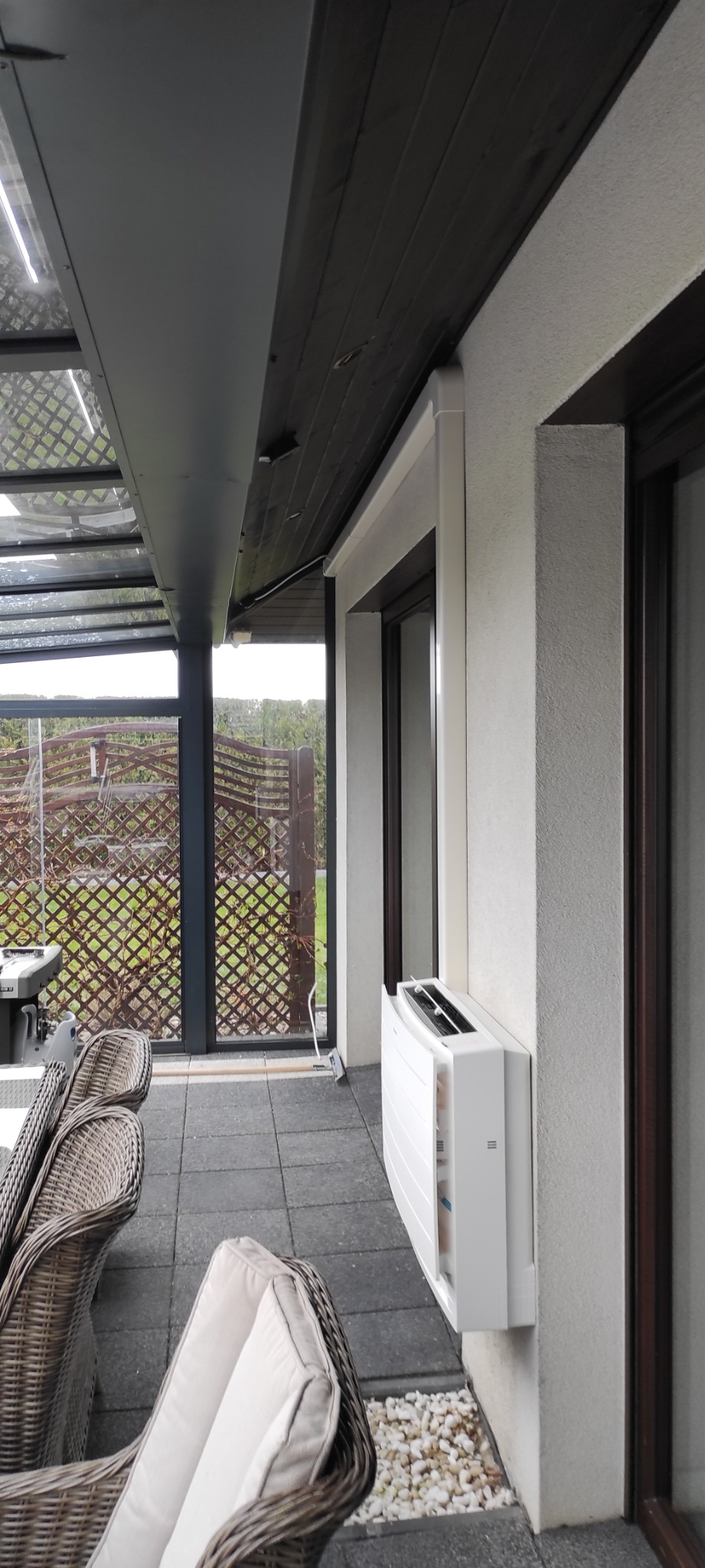 Biały klimatyzator przypodłogowy zamontowany na ścianie werandy, z widocznymi krzesłami z rattanu i przeszklonym dachem.