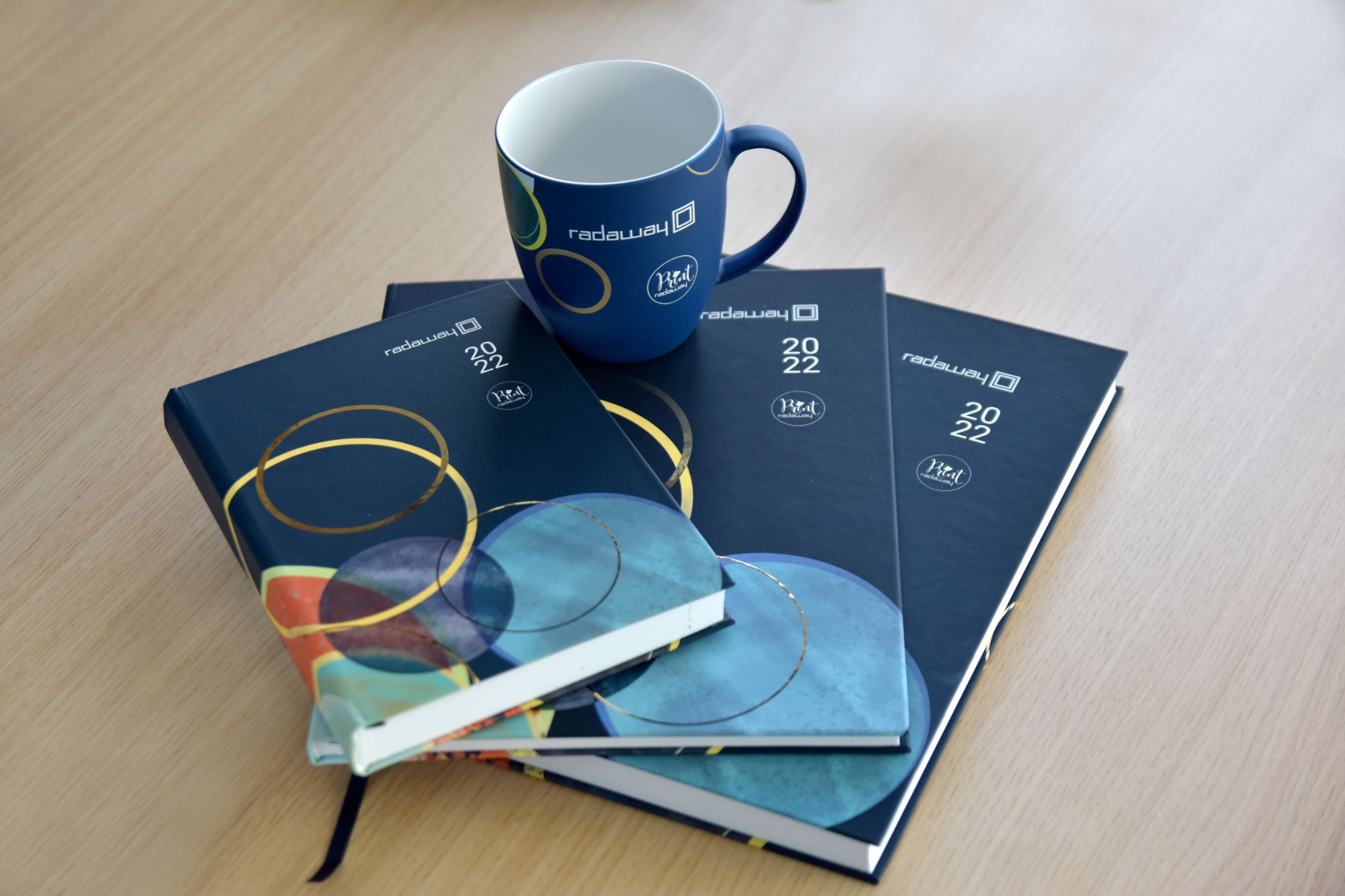 Zestaw firmowy ze Szczytnik: trzy granatowe kalendarze z logo i abstrakcyjnym wzorem oraz dopasowany kubek z logo, ułożone na drewnianym stole.