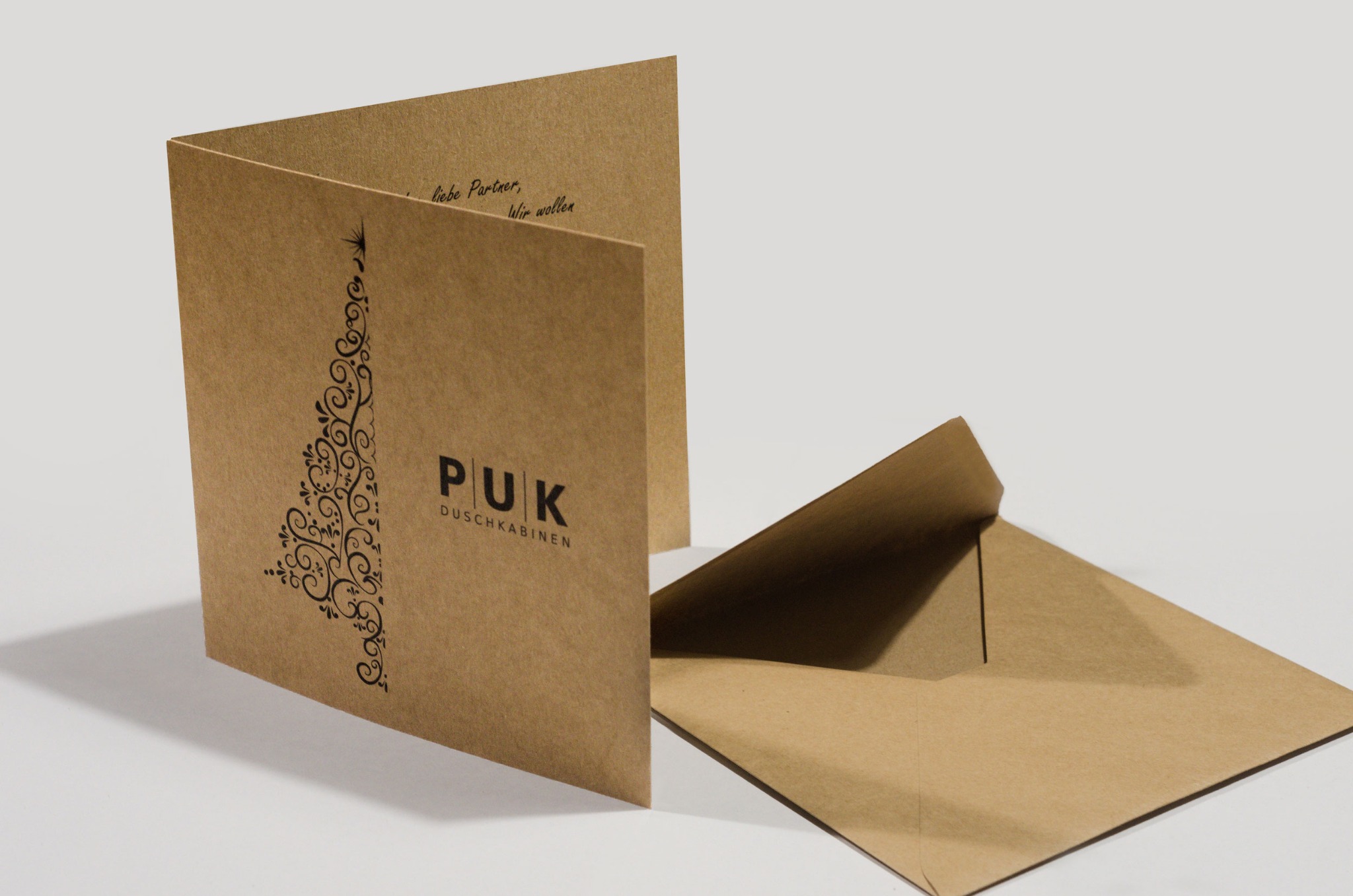 Otwarta, brązowa kartka z ozdobnym motywem choinki i logo PUK DUSCHKABINEN, obok brązowa koperta na białym tle.