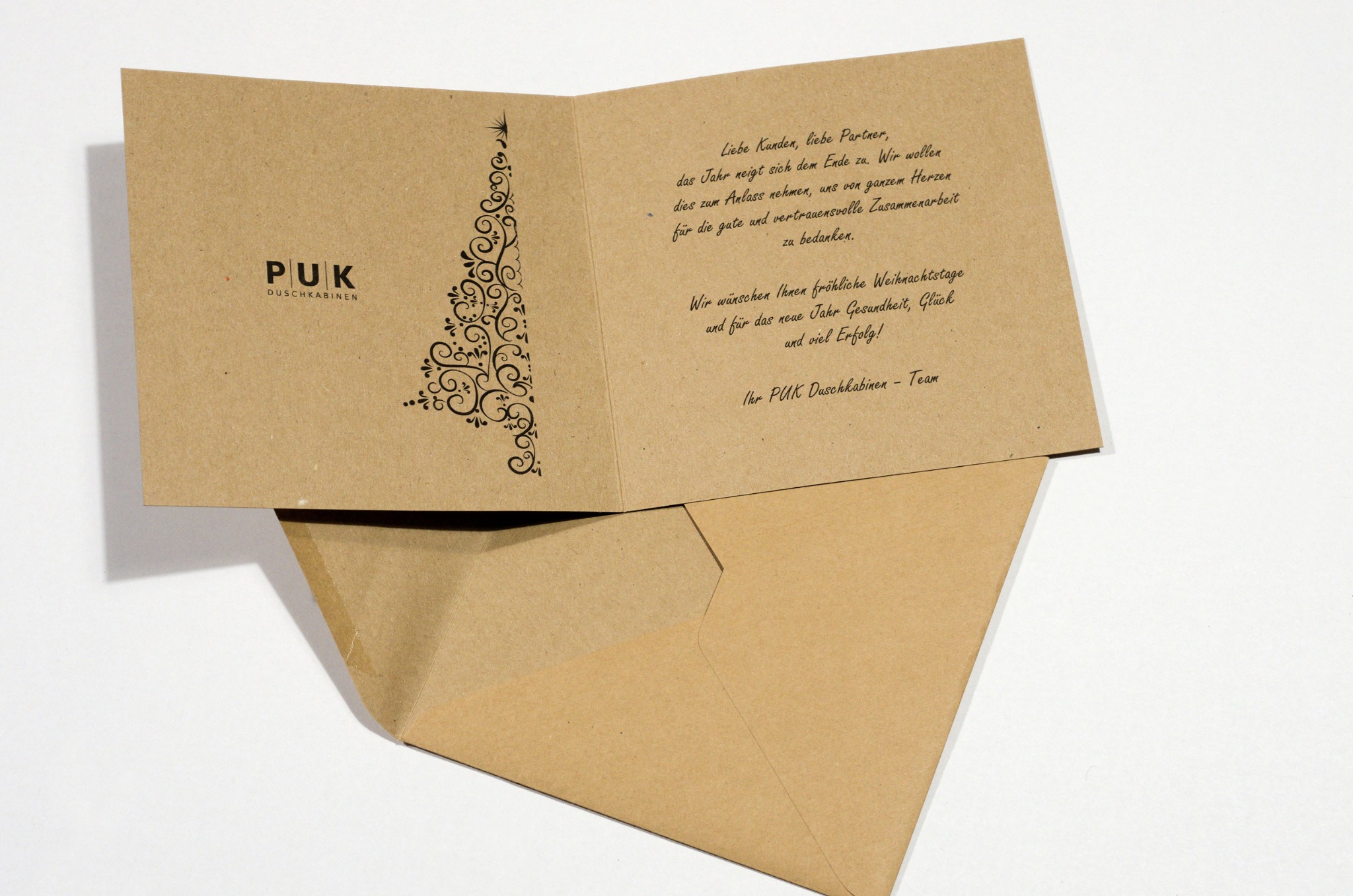 Otwarta kartka świąteczna z brązowego papieru z logo firmy PUK Duschkabinen i dekoracyjnym motywem choinki, leżąca na kopercie w odcieniu brązu.