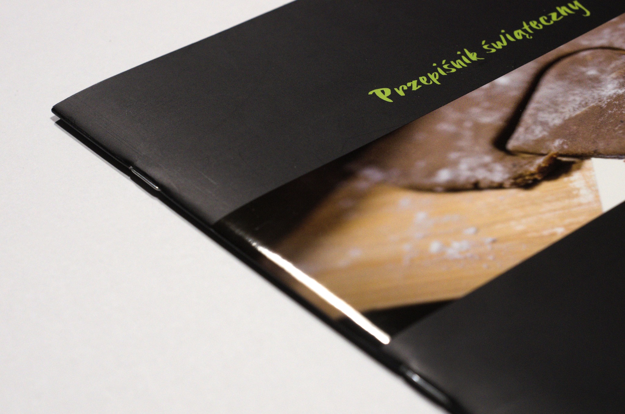 Otwarta broszura z czarną okładką i zielonym napisem 'Przepiśnik świąteczny', prezentująca zdjęcie ciasteczek piernikowych wycinanych z ciasta na drewnianej desce.