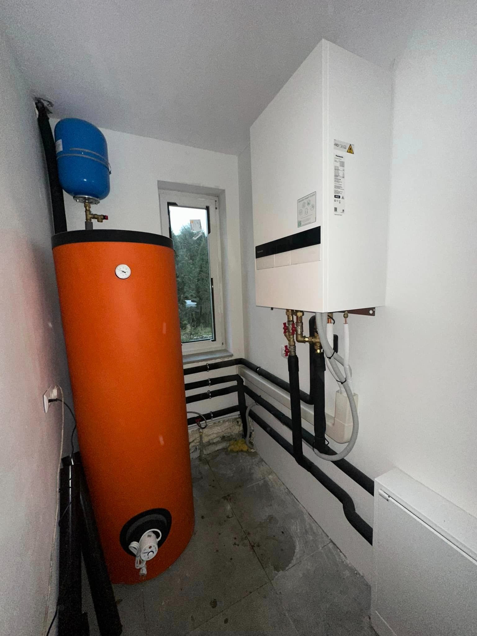Pomieszczenie z zainstalowaną białą jednostką pompy ciepła wiszącą na ścianie, obok stoi pomarańczowy zasobnik na wodę z niebieskim zbiornikiem wyrównawczym, widoczne rury instalacyjne.