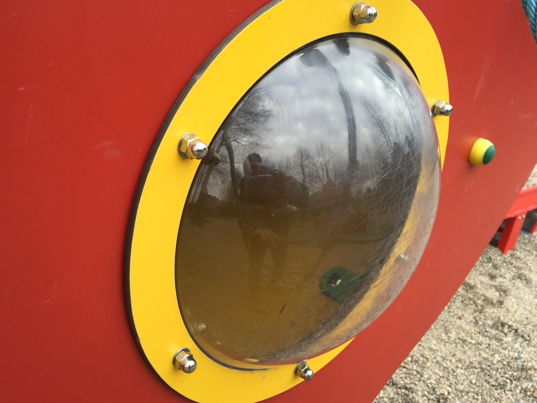 Czerwona ściana z żółtą ramą i przezroczystym, wypukłym okienkiem, w którym odbija się sylwetka osoby i drzewa.