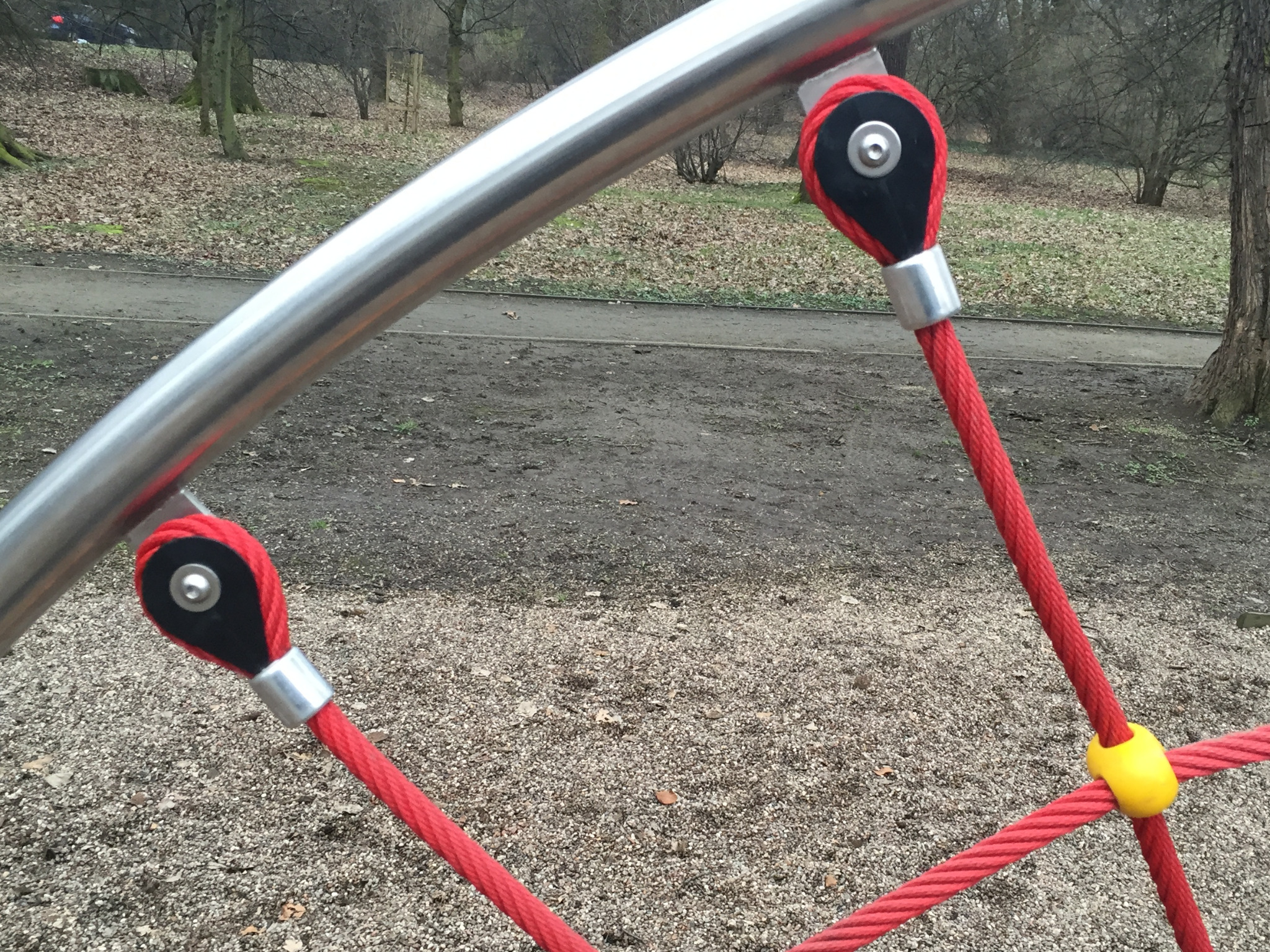 Element konstrukcyjny linowej piramidy w parku: stalowa rura, czerwone liny z czarnymi kauszami i żółtym łącznikiem na żwirowym podłożu.