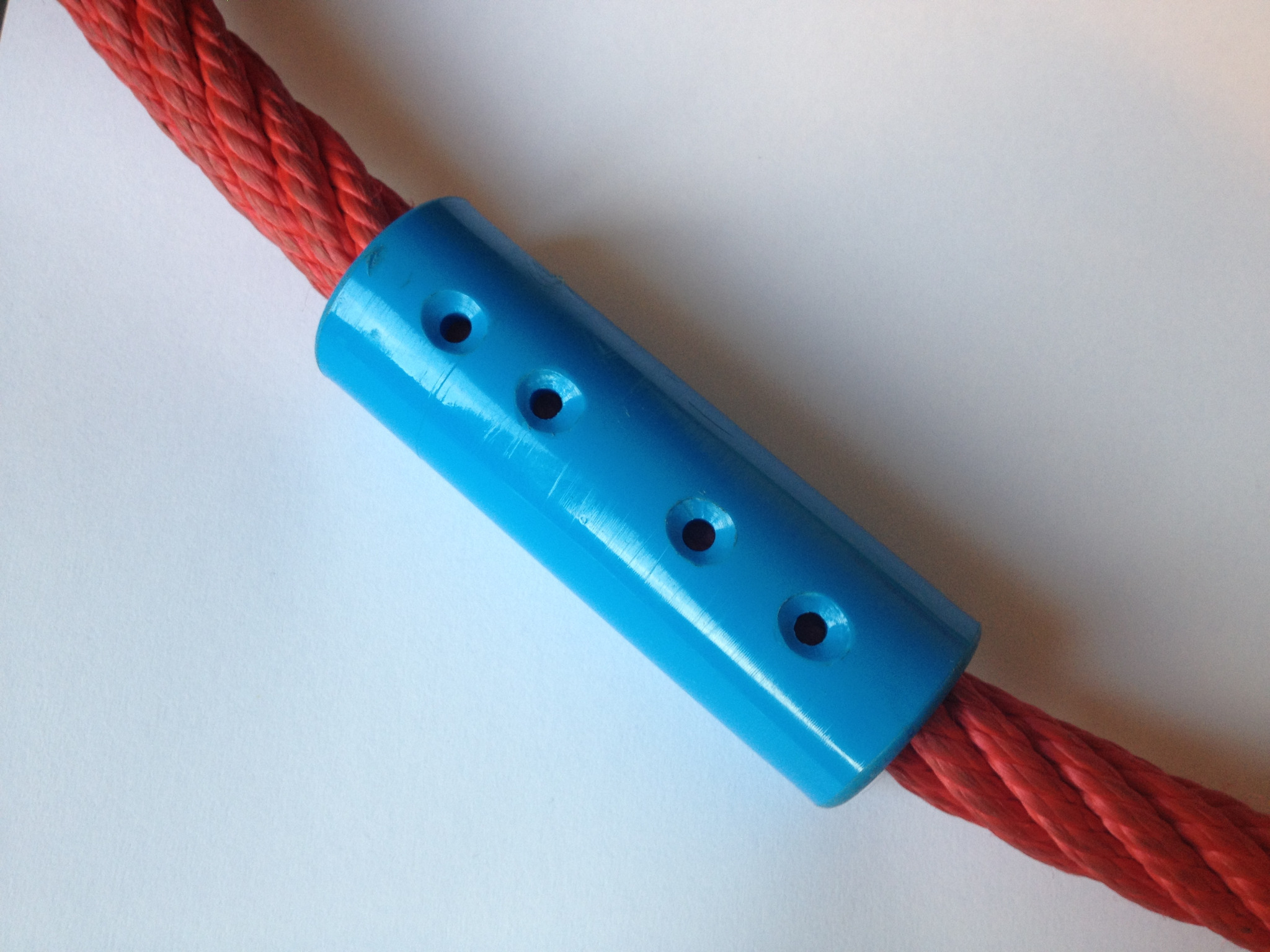 Czerwona lina zabezpieczona niebieską, plastikową osłoną z otworami na białym tle, element konstrukcyjny wyposażenia placu zabaw.