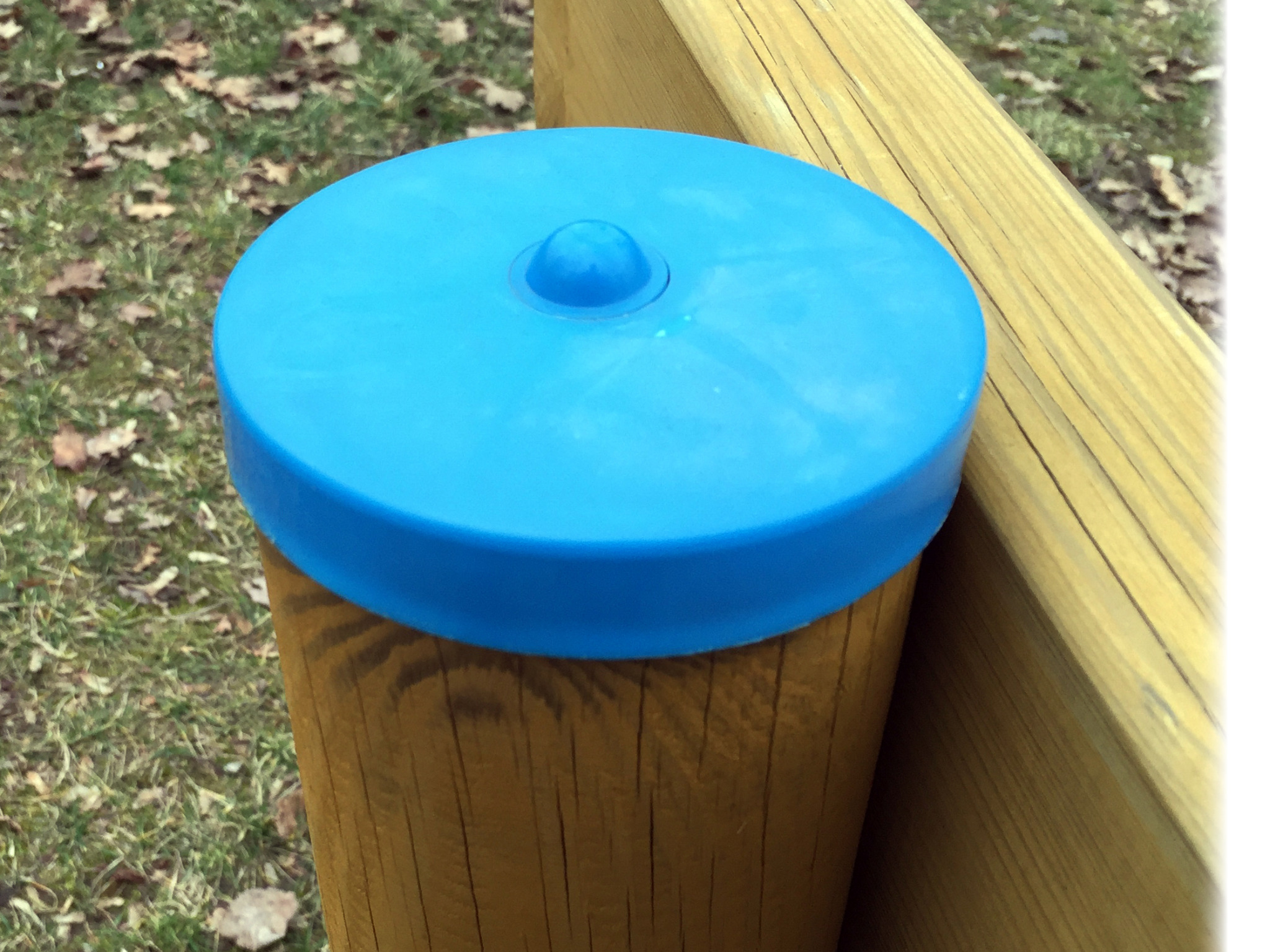 Niebieska, plastikowa nakładka z wypustką na drewnianym słupku konstrukcji, prawdopodobnie elementu wyposażenia placu zabaw, z drewnianą barierką w tle.