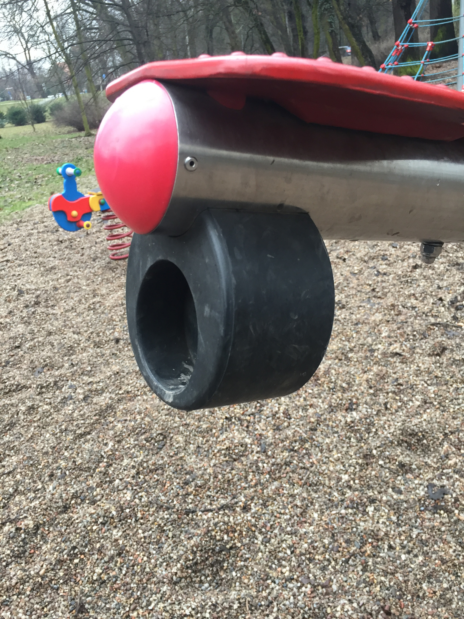 Fragment konstrukcji urządzenia na placu zabaw w kształcie samolotu z czerwonym nosem i czarnym kołem, na żwirowym podłożu.