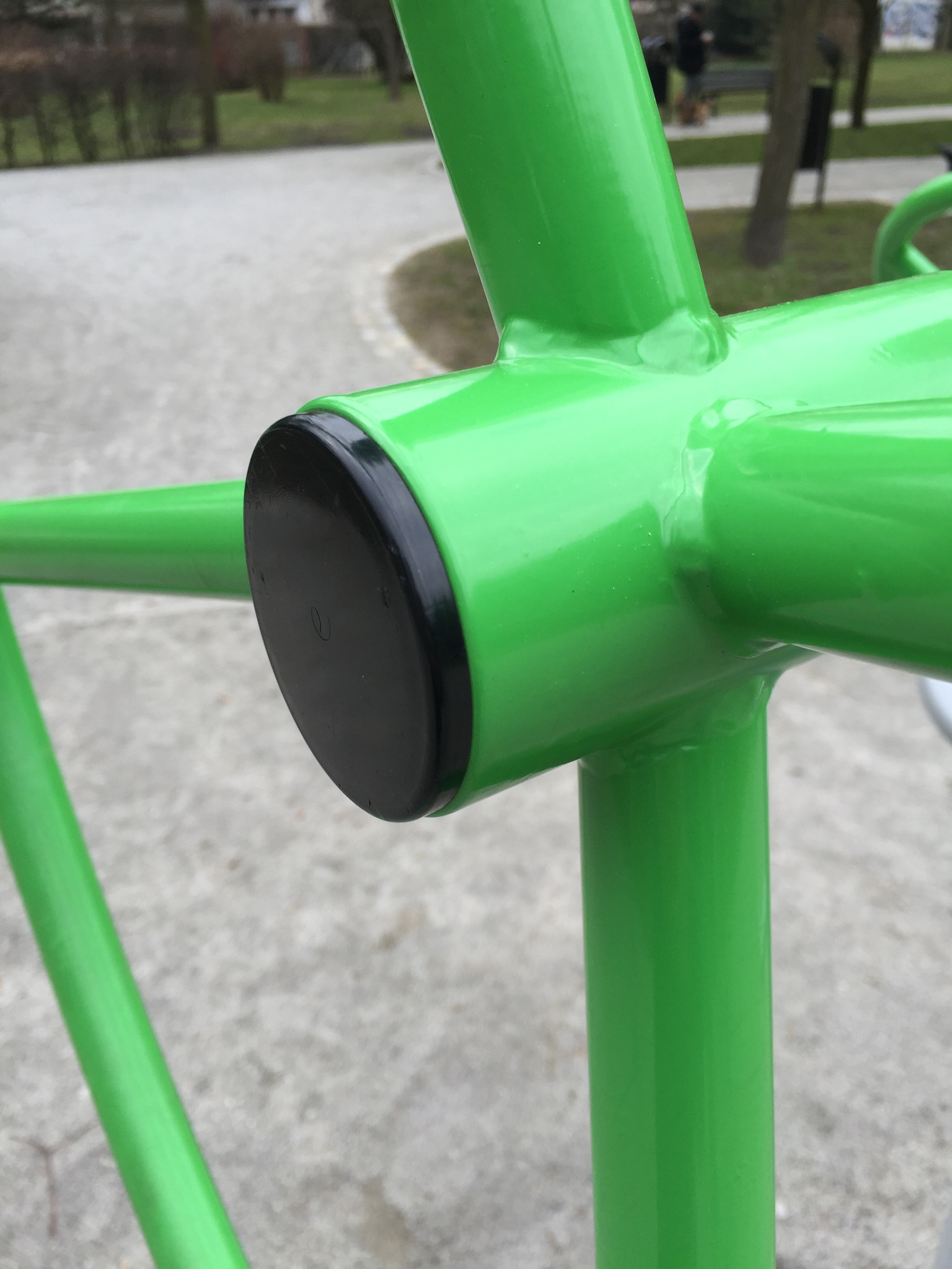 Fragment zielonej metalowej konstrukcji z czarnym zaślepkiem na tle parku.