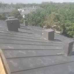 Szary blaszany dach z kominkami wentylacyjnymi na tle zabudowy i zieleni, widoczne ślady po czyszczeniu lub montażu.