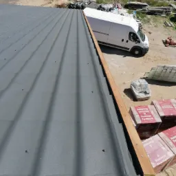 Montaż szarej blachodachówki na drewnianej łacie, z widocznymi paczkami materiałów budowlanych Wienerberger Porotherm i białym samochodem dostawczym w tle.