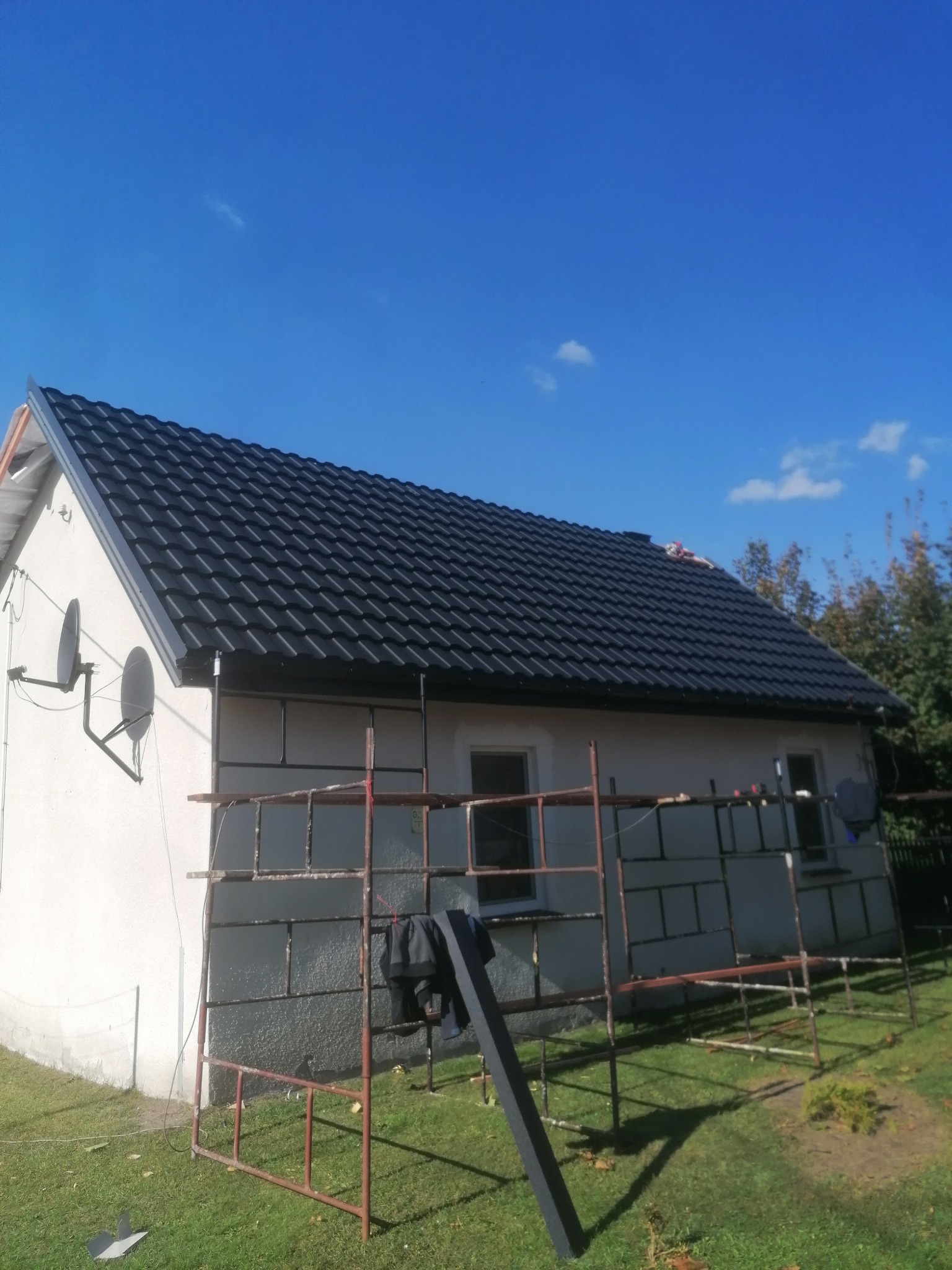 Dom z nowym, czarnym dachem z blachodachówki, widoczne rusztowania i anteny satelitarne na ścianie, błękitne niebo w tle.