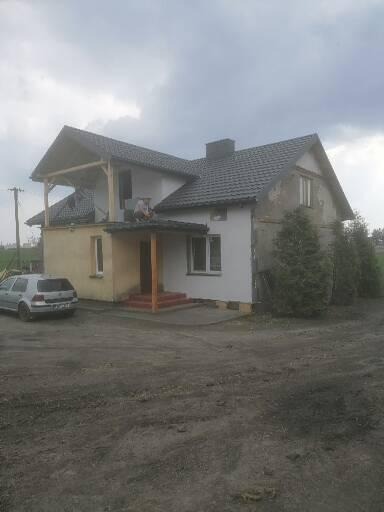 Dom jednorodzinny z szarą dachówką, częściowo ocieplony, z widocznym fragmentem konstrukcji drewnianej i pracownikiem na dachu werandy.