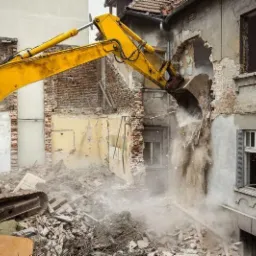 Żółta koparka wyburza fragment ściany budynku, z gruzem i pyłem w powietrzu, odsłaniając ceglaną konstrukcję wewnątrz.