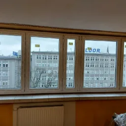 Nowe, białe okno PCV z widokiem na miejski budynek z szarym niebem w tle, widoczne ślady po montażu na ścianie.