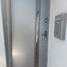 Nowoczesne, szare drzwi wejściowe z wąskim przeszkleniem, zamontowane w szarej ścianie obok skrzynki na listy z numerem 19.