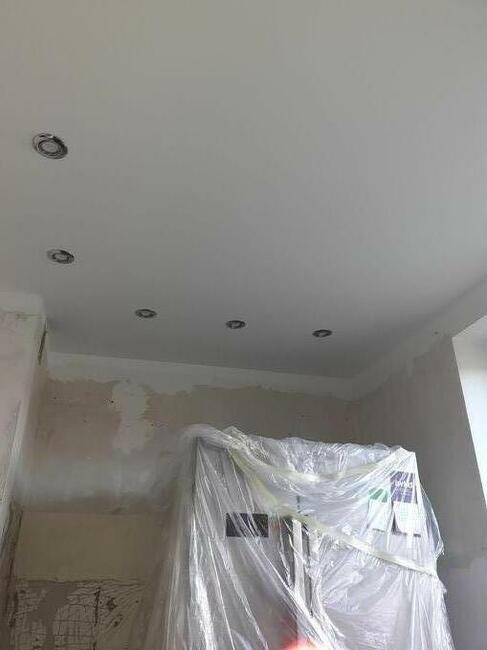 Biały sufit z zamontowanymi punktowymi oprawami oświetleniowymi; ściany w trakcie przygotowania, z widocznymi śladami szpachlowania i zabezpieczone folią meble w tle.