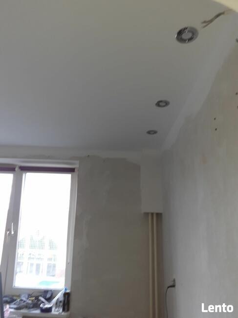 Biały sufit z zamontowanymi okrągłymi lampami halogenowymi, ściany w trakcie wykończenia, widoczne ślady po gładzi, okno z białymi ramami.