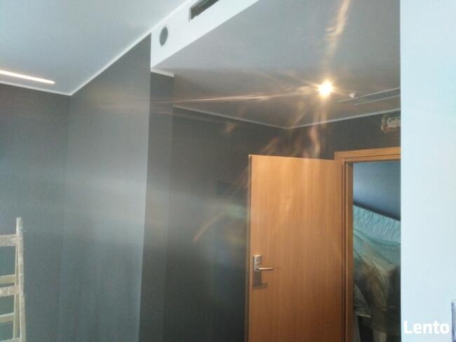 Świeżo pomalowane na szaro ściany w pokoju z widoczną drabiną malarską i otwartymi drzwiami do kolejnego pomieszczenia, gdzie widać zabezpieczoną folią przestrzeń.