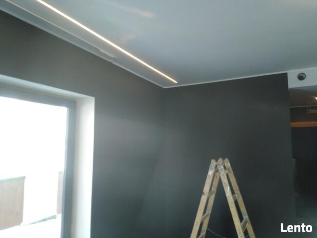 Szare wnętrze z ukrytym oświetleniem LED w suficie i drabiną opartą o ścianę, widok na okno po lewej stronie.