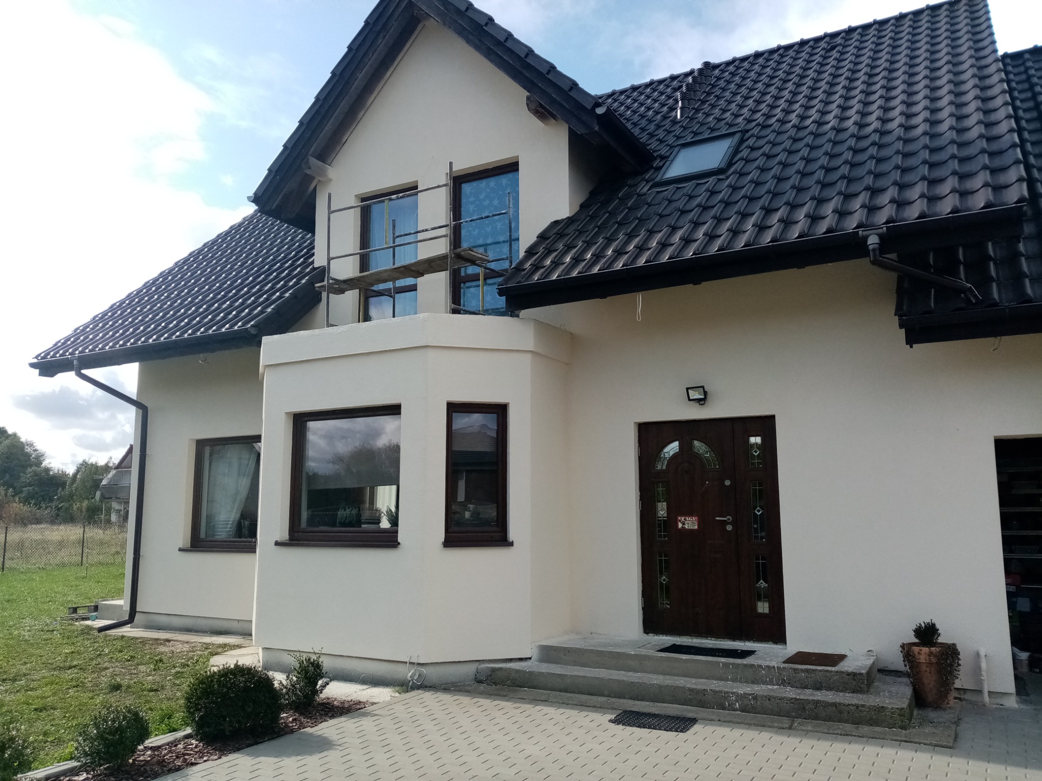 Dwupiętrowy dom z białą elewacją, czarnym dachem z dachówki i brązowymi oknami. Widoczne rusztowanie przy oknach na piętrze, sugerujące prace wykończeniowe. Jasne niebo w tle.
