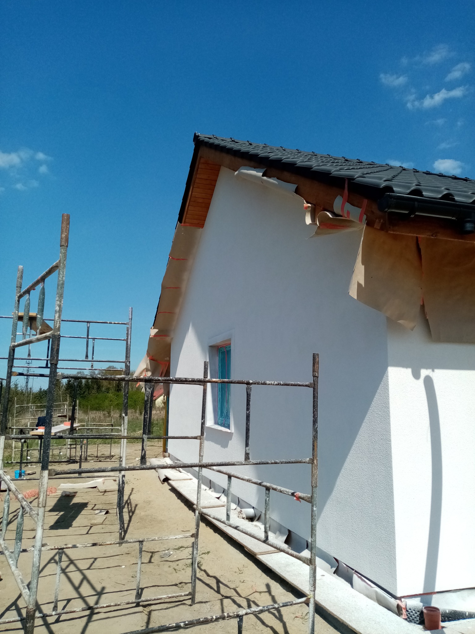 Boczny widok domu z nową, białą elewacją o wyraźnej fakturze, z widocznymi rusztowaniami, zabezpieczeniami okien i fragmentami taśmy malarskiej na dachu, w słoneczny dzień.