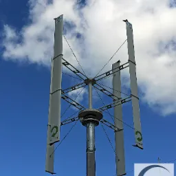 Pionowa turbina wiatrowa z logo Ecorote na tle błękitnego nieba z chmurami, widok od dołu na zainstalowaną konstrukcję.