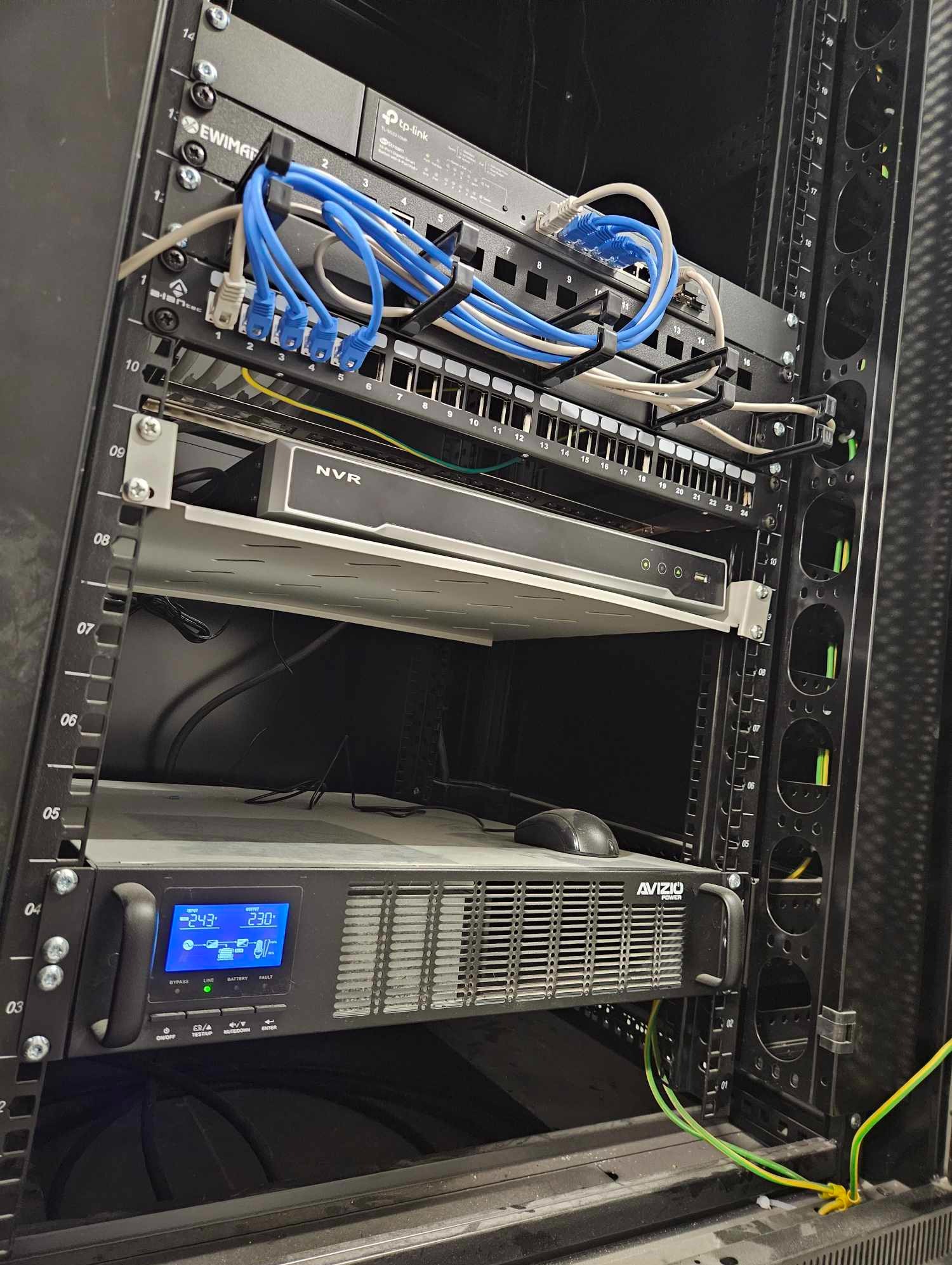 Szafa rack z zainstalowanym sprzętem sieciowym: switch, rejestrator NVR i zasilacz awaryjny UPS marki Avizio, okablowanie sieciowe z niebieskimi kablami Ethernet, przewody uziemiające w kolorze...