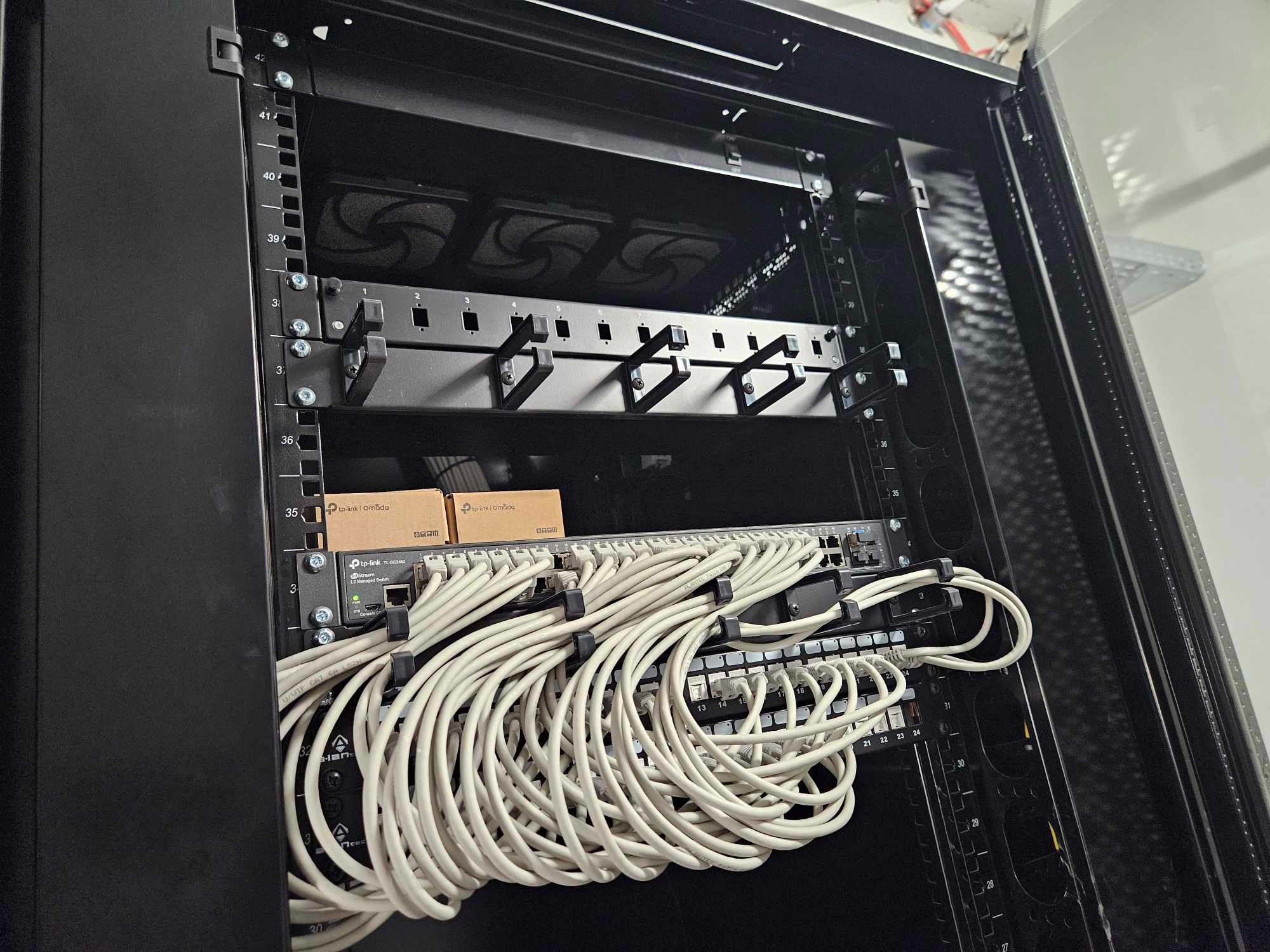 Szafa serwerowa z zainstalowanym switchem TP-Link Omada, uporządkowane białe kable sieciowe podłączone do portów, widoczne oznaczenia numerów na obudowie szafy.