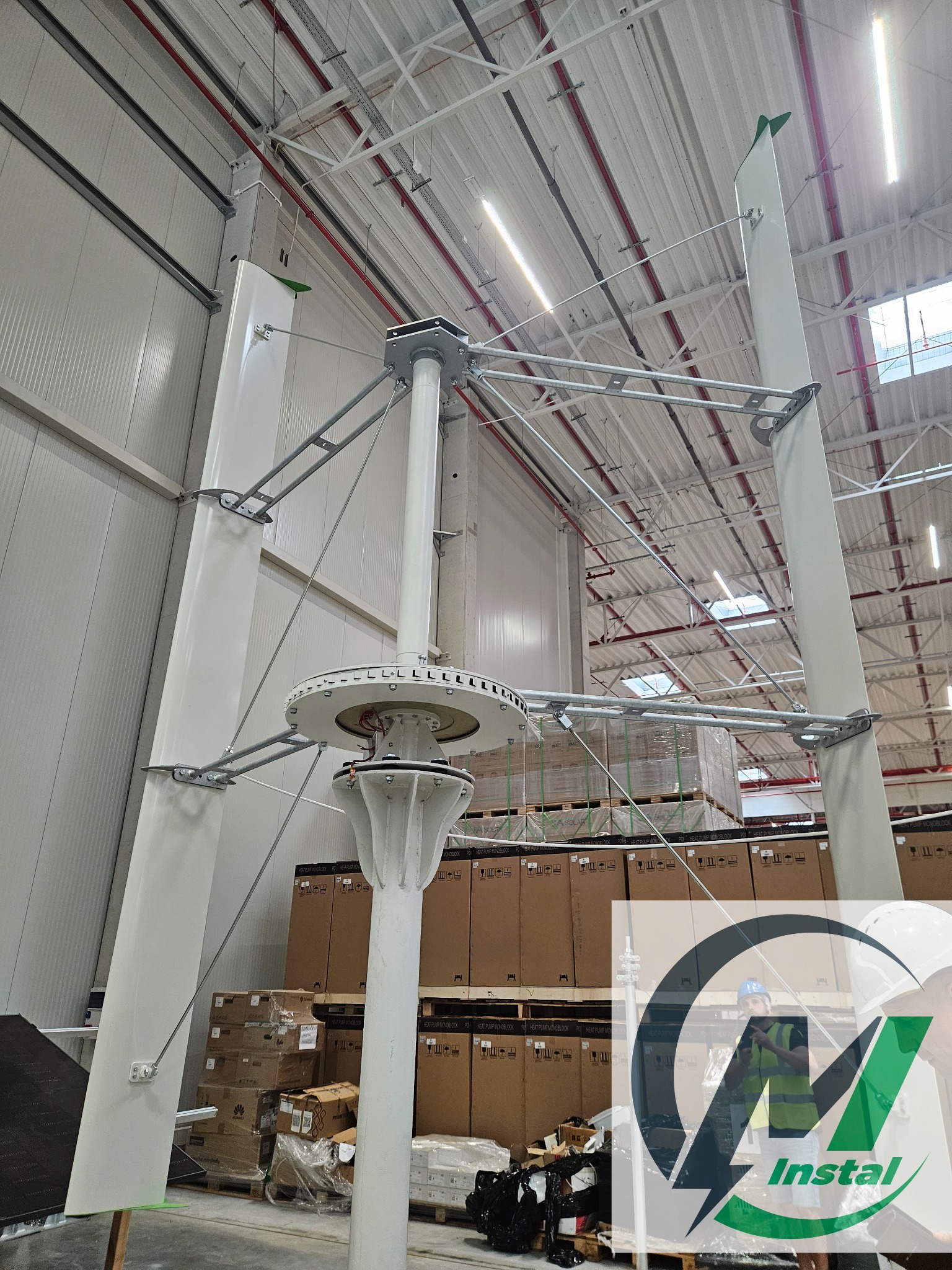 Wertykalna turbina wiatrowa typu Darrieusa, konstrukcja z białymi łopatami i stalowymi wspornikami, widoczna w hali magazynowej z kartonami w tle, logo firmy częściowo zasłania widok.