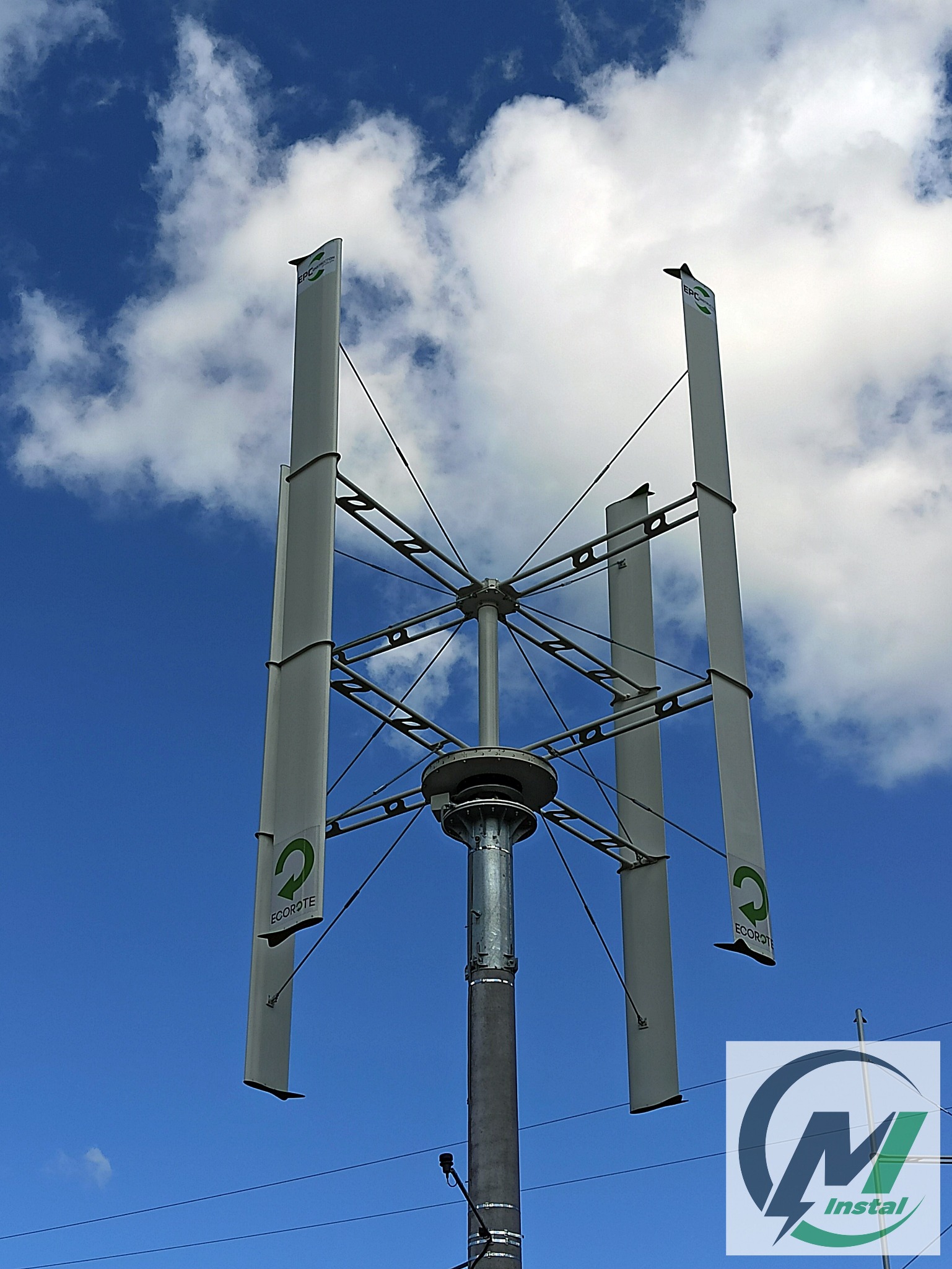 Pionowa turbina wiatrowa z logo Ecorote na tle błękitnego nieba z chmurami, widok od dołu na zainstalowaną konstrukcję.