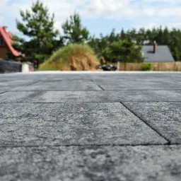 Szare płyty tarasowe ułożone z minimalnymi fugami, w tle dom z białymi elementami architektonicznymi i zieleń drzew, widok z niskiej perspektywy podkreśla fakturę kamienia.