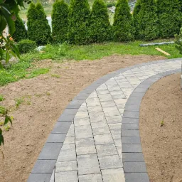 Zakrzywiony chodnik z jasnoszarej kostki brukowej obramowany ciemniejszą kostką, prowadzący przez trawnik z tujami w tle.