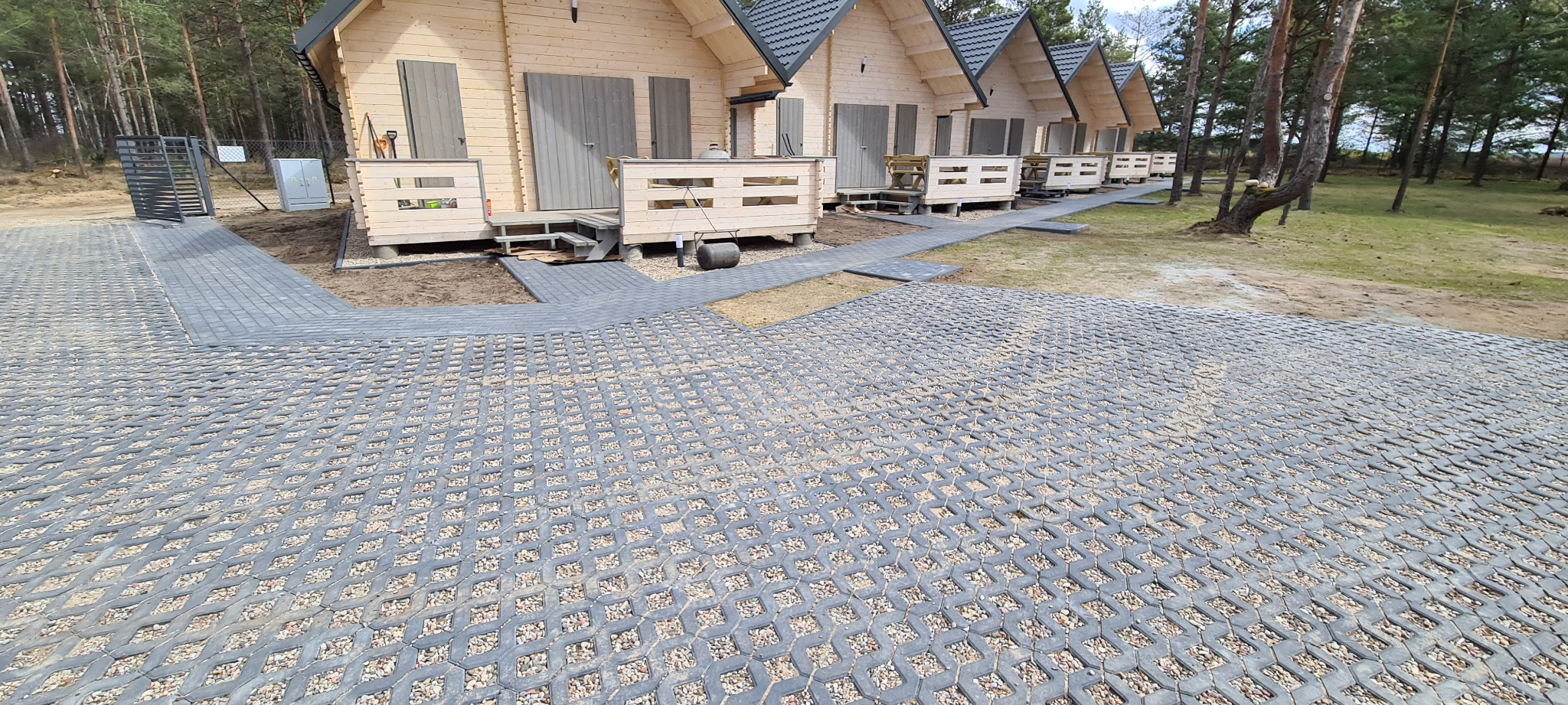 Szara ażurowa nawierzchnia z wypełnieniem żwirowym prowadząca do drewnianych domków letniskowych z szarą stolarką, widok na bramkę wejściową.