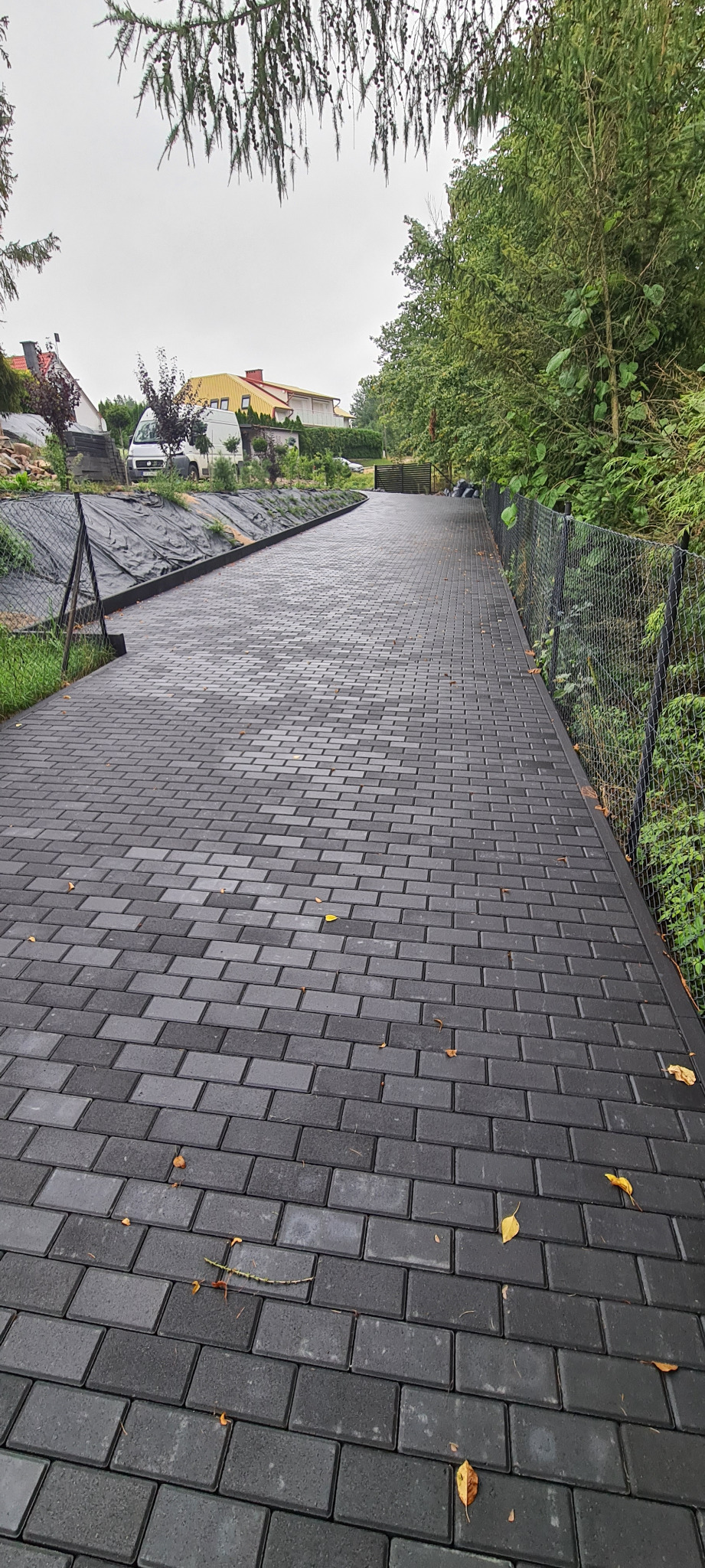 Długa, nowa, szara ścieżka z kostki brukowej prowadząca wzdłuż ogrodzenia z siatki, z widocznymi domami w tle.