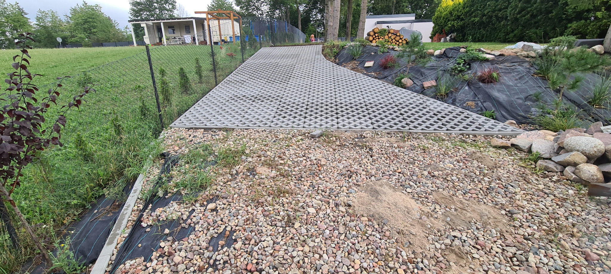 Ułożona nawierzchnia z ażurowych płyt betonowych na podjeździe, widoczne kamienie ozdobne i agrowłóknina na skarpie, w tle dom jednorodzinny i drzewa.