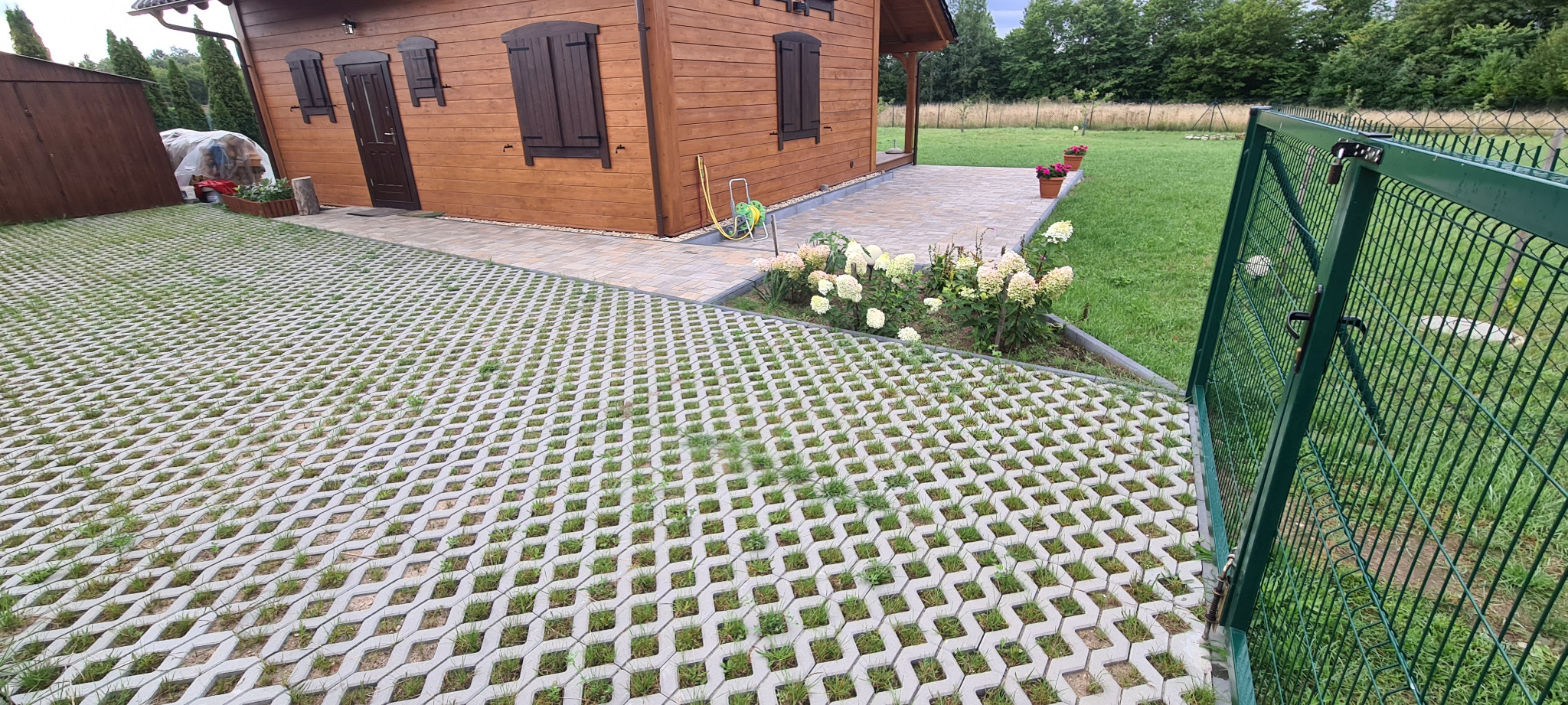 Ażurowy podjazd z kostki betonowej z widoczną trawą, prowadzący do drewnianego domu z brązowymi okiennicami i furtki ogrodzeniowej.