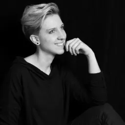 Portret uśmiechniętej kobiety z krótkimi blond włosami, w czarnej bluzie z kapturem, z kolczykiem w uchu i brwi, zamyślona, trzymająca dłoń przy ustach, na czarnym tle.